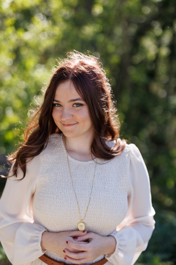 Ava posing for her high school senior portrait in Hudson, Wisconsin, wearing a light blouse and gold necklace, surrounded by greenery.