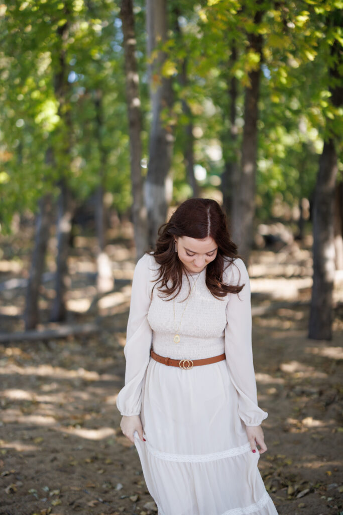 Ava in a flowing white dress and brown belt, looking down amidst trees in Hudson, WI during her high school senior photography session.