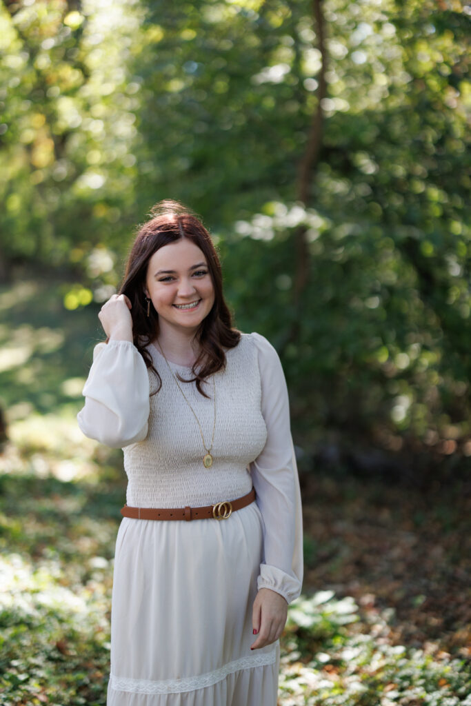 Ava smiling in a white dress and brown belt, celebrating her high school graduation in a lush, green outdoor setting in Hudson, Wisconsin.