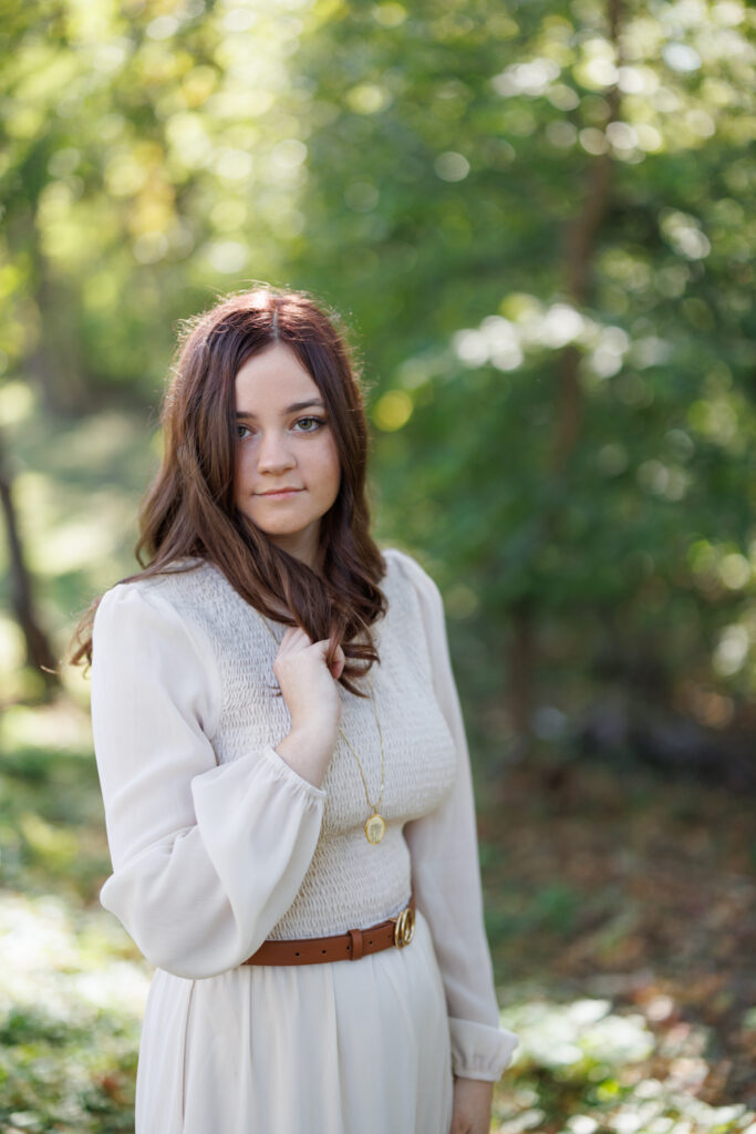 Ava in a light-colored dress and necklace, posing outdoors amidst greenery during her high school senior photography session in Hudson, Wisconsin.