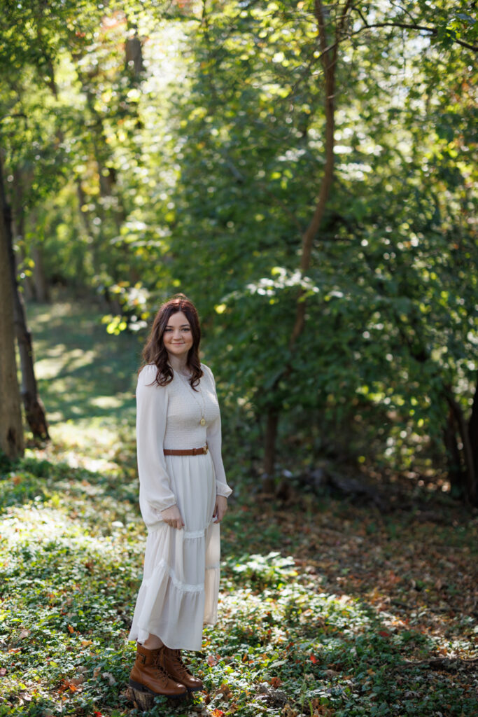 Ava in a white dress and brown boots standing in a lush green forest during her high school senior photography session in Hudson, Wisconsin.