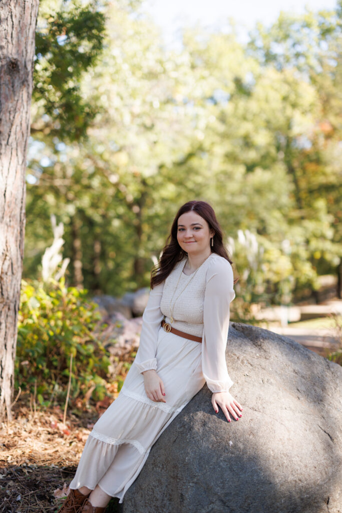 Ava in a white dress sitting on a rock surrounded by greenery, celebrating her high school graduation in Hudson, Wisconsin.
