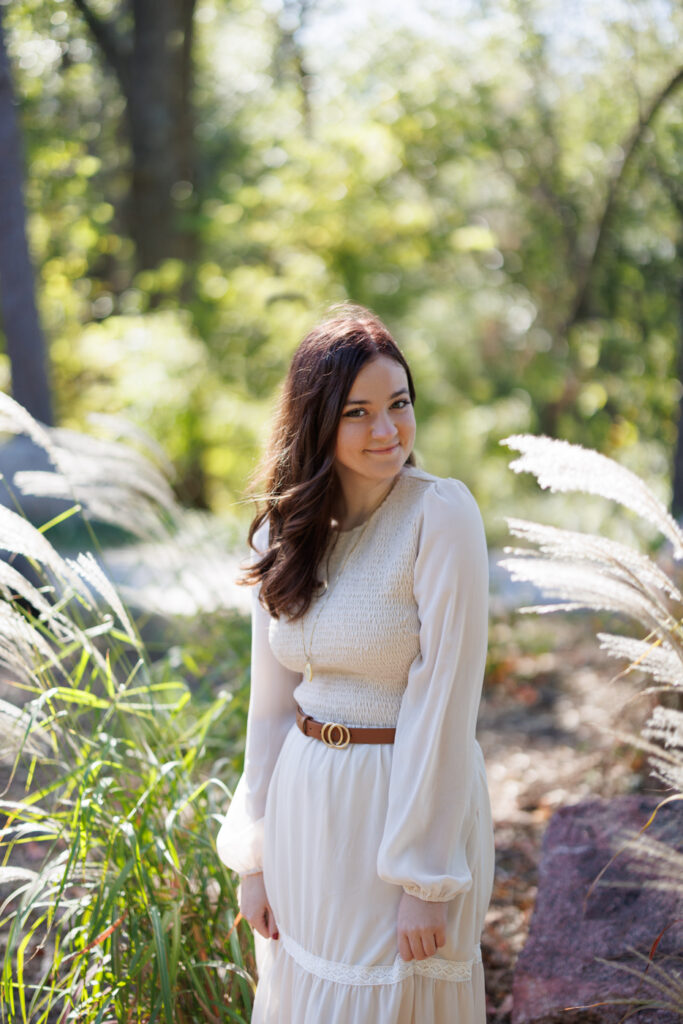 Ava posing in a flowing white dress with a belt, surrounded by greenery during her senior portrait session in Hudson, Wisconsin.