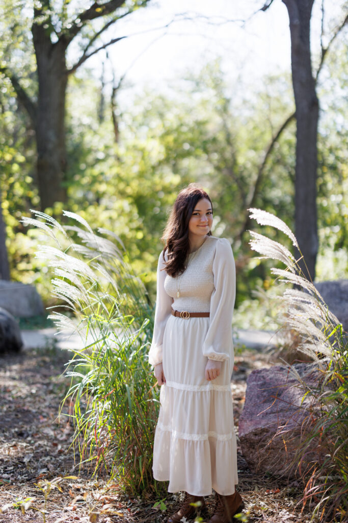 Ava in a flowing white dress and brown boots, smiling among tall grasses and trees during her senior portrait session in Hudson, Wisconsin.