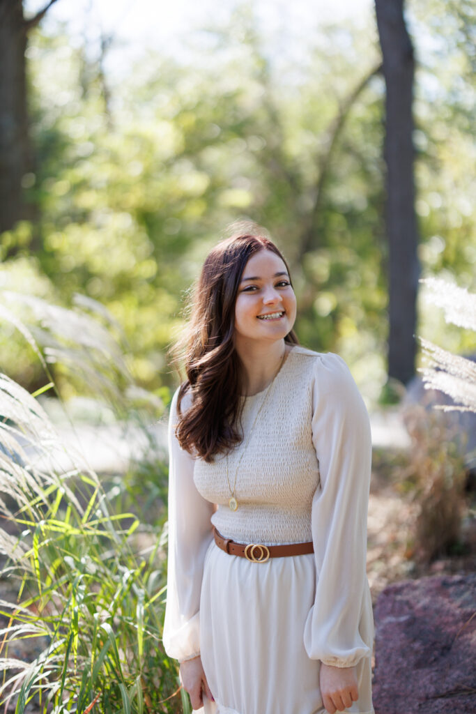 Ava smiling in a white dress with a belt, surrounded by greenery during her senior photo session in Hudson, Wisconsin.