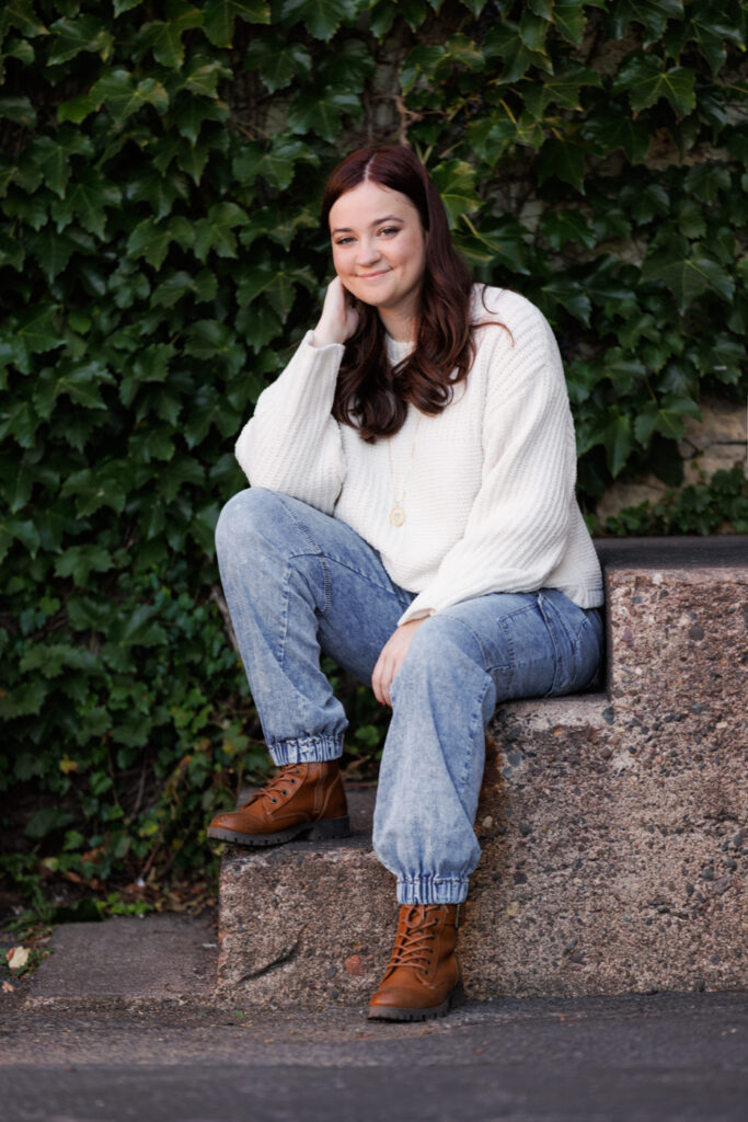Ava sitting on a stone step in Hudson, WI, wearing a cozy white sweater and blue jeans, smiling against a backdrop of green ivy, celebrating her high school graduation.