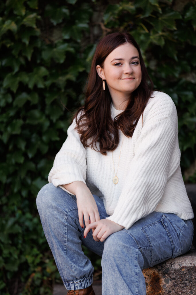 Ava sitting on a stone, wearing a cozy white sweater and denim jeans, smiling against a backdrop of green ivy, celebrating her high school senior session in Hudson, Wisconsin.