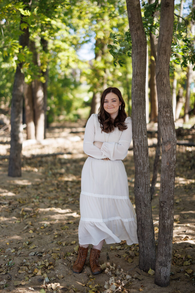 Ava posing in a white dress and brown boots among trees, celebrating her high school senior session in Hudson, Wisconsin.