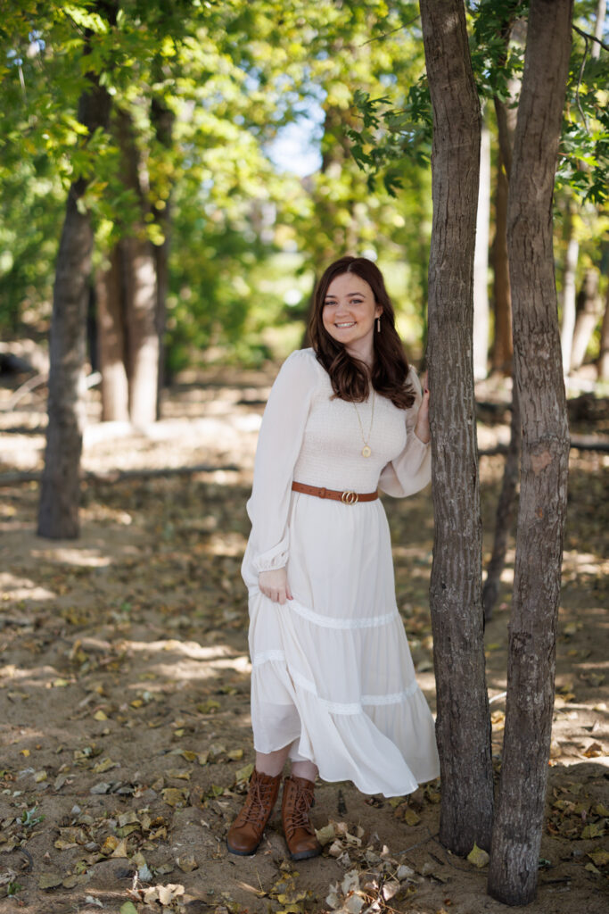 Ava in a flowing white dress and brown boots, posing beside a tree in a wooded area during her high school senior portrait session in Hudson, Wisconsin.