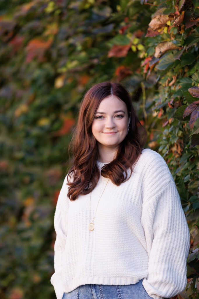 Ava smiling in a cozy white sweater against a vibrant green and autumn-colored backdrop, celebrating her high school graduation in Hudson, WI.