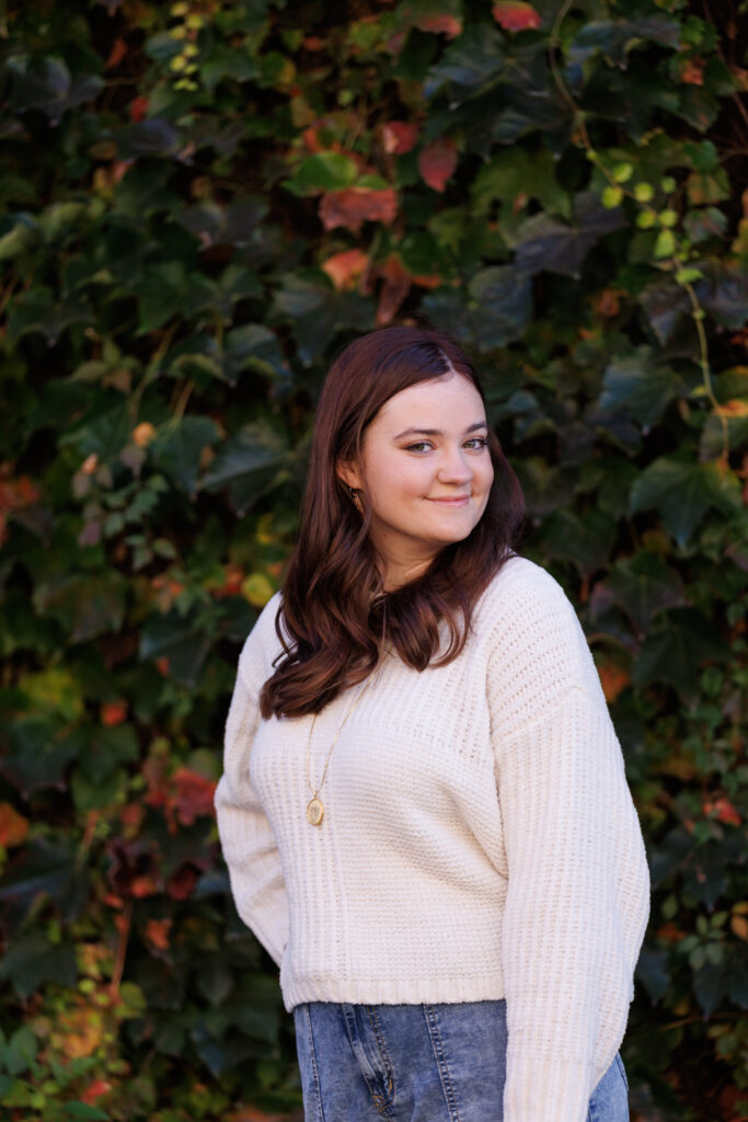 Ava smiling in a cozy white sweater against a backdrop of vibrant green and red ivy, celebrating her high school senior portrait session in Hudson, WI.