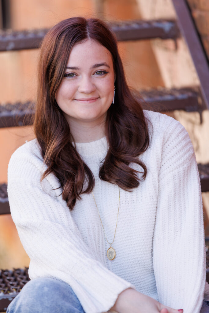 Ava smiling in a cozy white sweater, seated on steps during her high school senior portrait session in Hudson, Wisconsin.