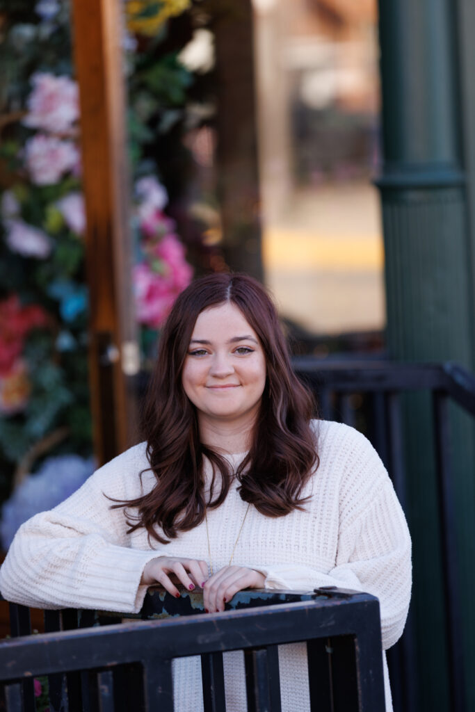 Ava smiling in a cozy cream sweater, leaning on a black railing with a vibrant floral backdrop, celebrating her high school graduation in Hudson, Wisconsin.