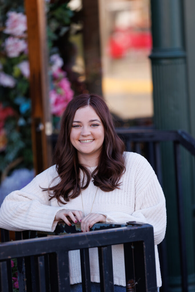 Ava smiling in cozy sweater, posing in Hudson, Wisconsin, surrounded by floral backdrop, celebrating her high school graduation session.