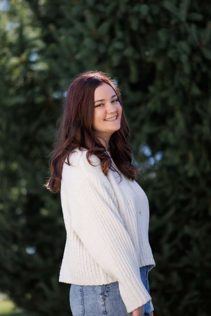 Ava smiling in a cozy white sweater, celebrating her high school graduation in Hudson, WI, surrounded by lush greenery.