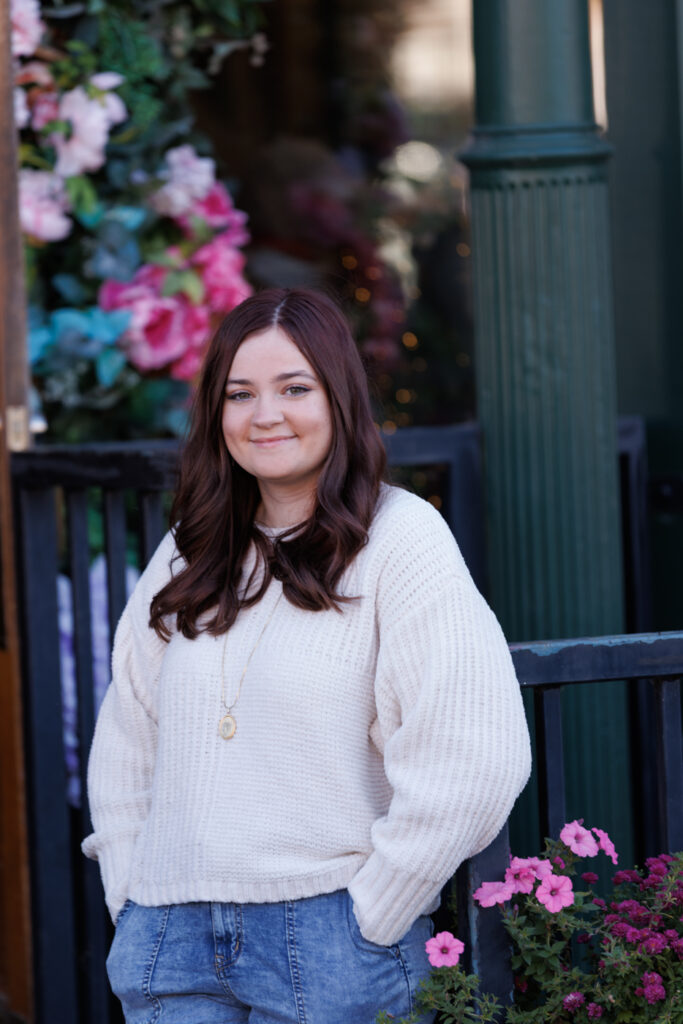 Ava posing in a cozy sweater with a smile, surrounded by colorful flowers in Hudson, WI, celebrating her high school senior portrait session.