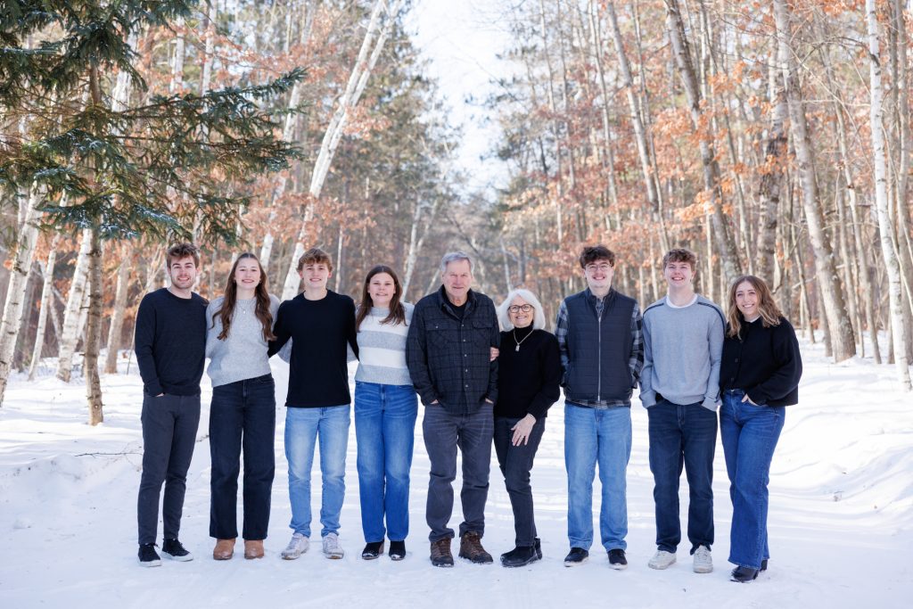 Winter family portrait in Spooner, Wisconsin, featuring eight family members in a snowy forest setting, showcasing warm clothing and smiles, highlighting family photography by Superior Sunset Studio.