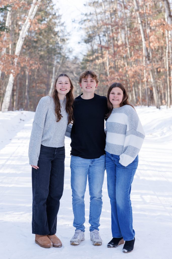 Winter family portrait in Spooner, Wisconsin, featuring three young adults smiling together in a snowy landscape, surrounded by trees with autumn leaves.