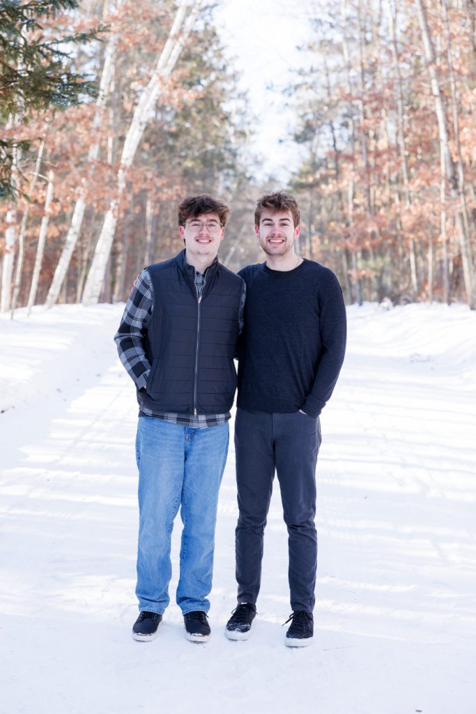 Two young men standing together in a snowy landscape in Spooner, Wisconsin, wearing winter clothing, smiling, surrounded by trees with autumn leaves.