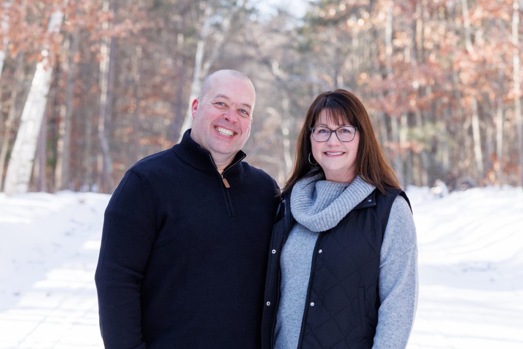 Smiling couple posing in snowy woods during winter family photography session in Spooner, Wisconsin.
