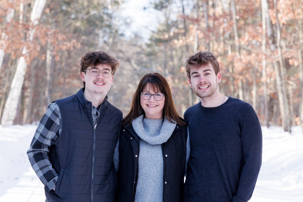 Winter family portrait in Spooner, Wisconsin, featuring a mother and two sons smiling together in a snowy forest setting.