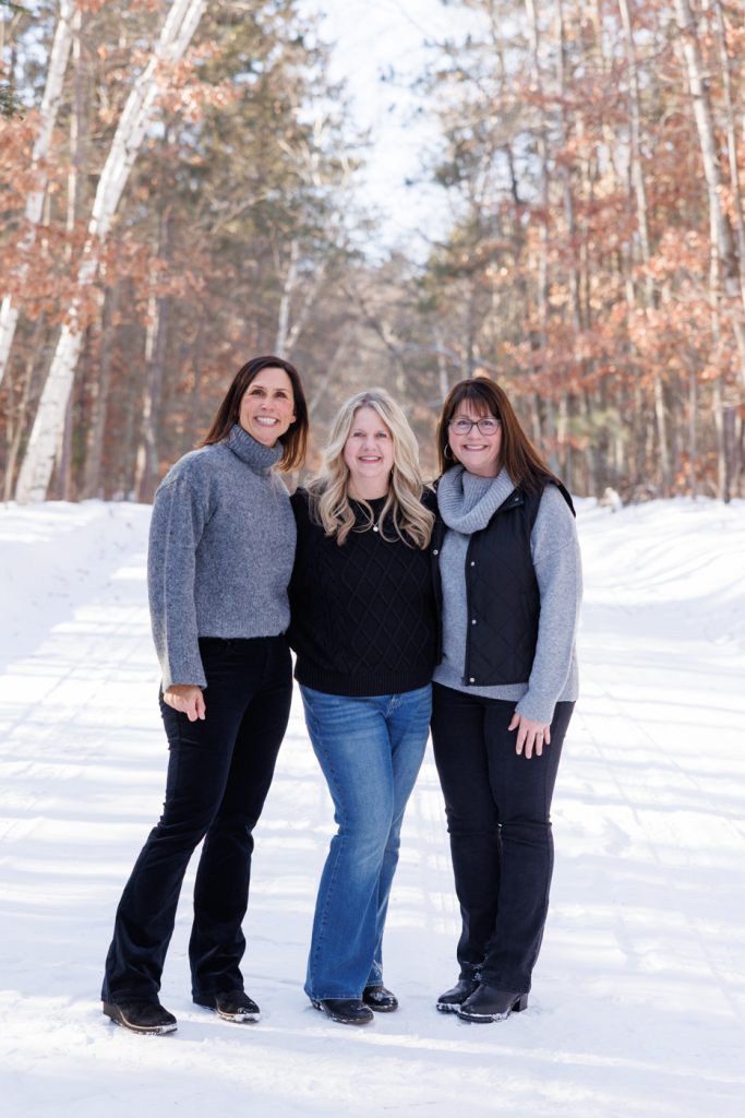 Three women smiling together in winter attire, standing in a snowy landscape with trees in Spooner, Wisconsin, showcasing a family photography session by Superior Sunset Studio.