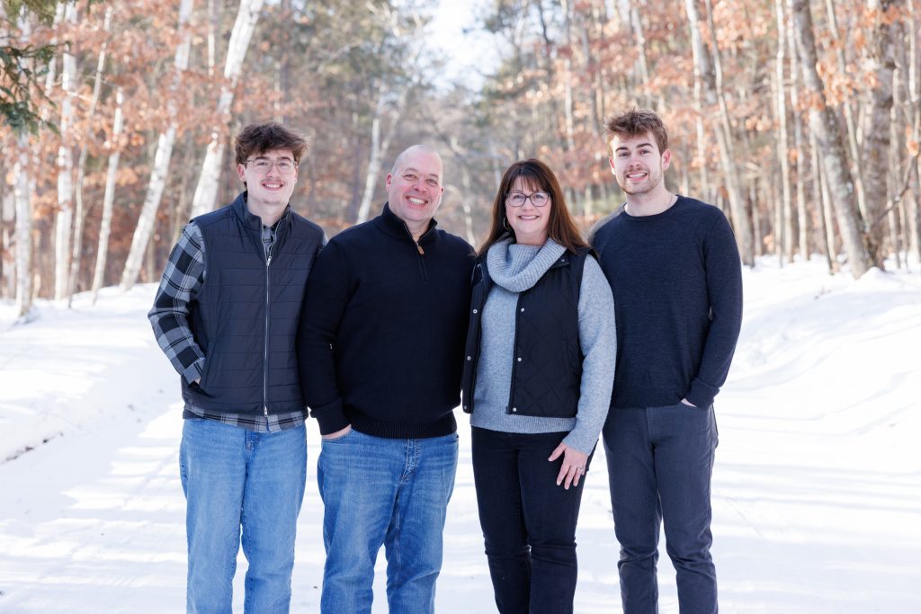 Winter family portrait in Spooner, Wisconsin, featuring four family members smiling together in a snowy forest setting.