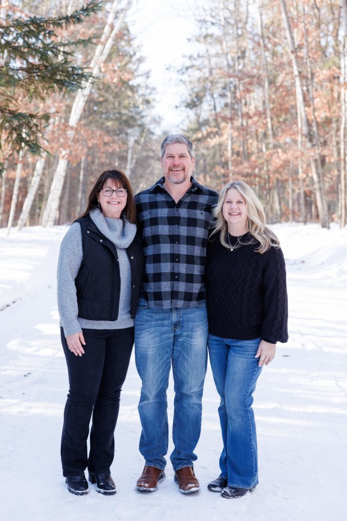Family portrait in winter setting, Spooner, Wisconsin, featuring three adults smiling, surrounded by snow and trees.