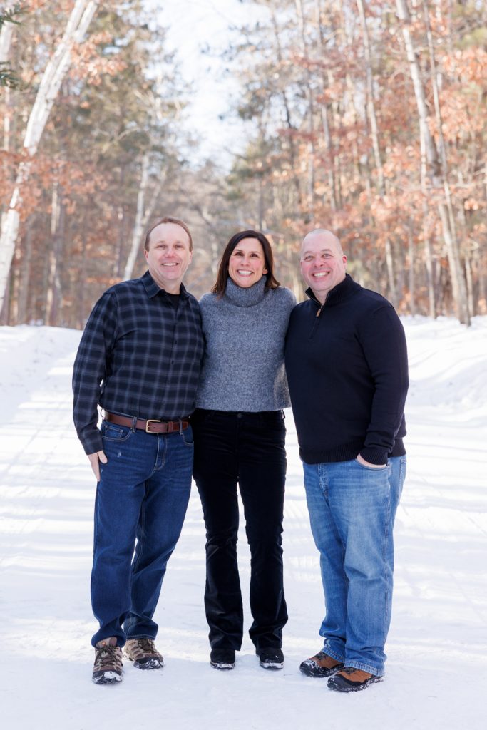 Winter family portrait in Spooner, Wisconsin, featuring three smiling adults dressed in cozy attire, surrounded by a snowy landscape and trees.