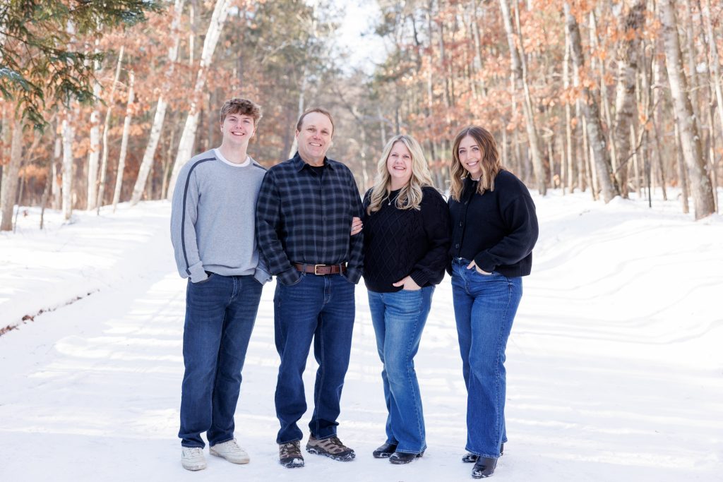 Winter family portrait in Spooner, Wisconsin, featuring four family members smiling in a snowy forest setting, showcasing Superior Sunset Studio's family photography session.