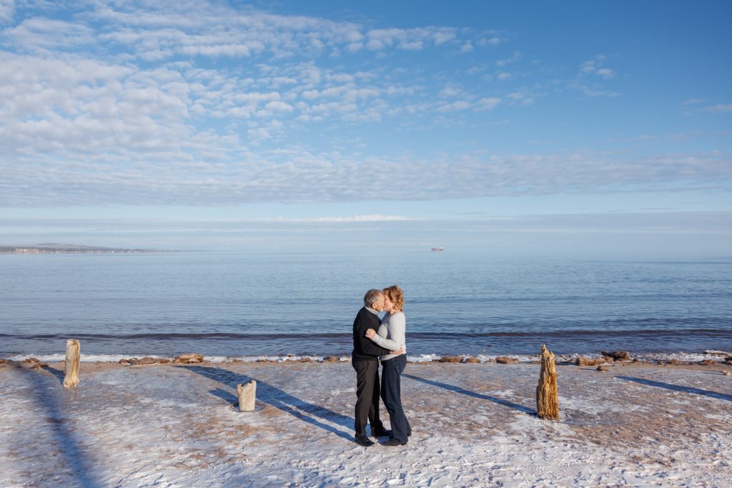 Couple embracing on a snowy beach in Duluth, Minnesota, with Lake Superior in the background and a clear blue sky.