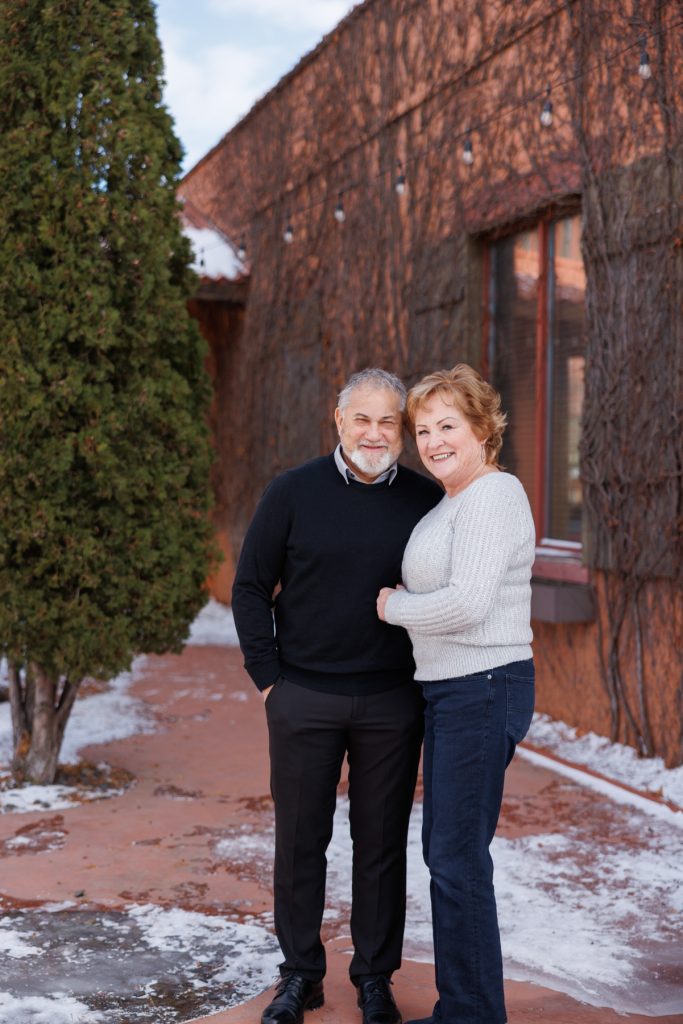Jane and Bruce smiling together in winter attire, standing on a snowy path in Duluth, Minnesota, with a rustic building and greenery in the background, celebrating their photography session.