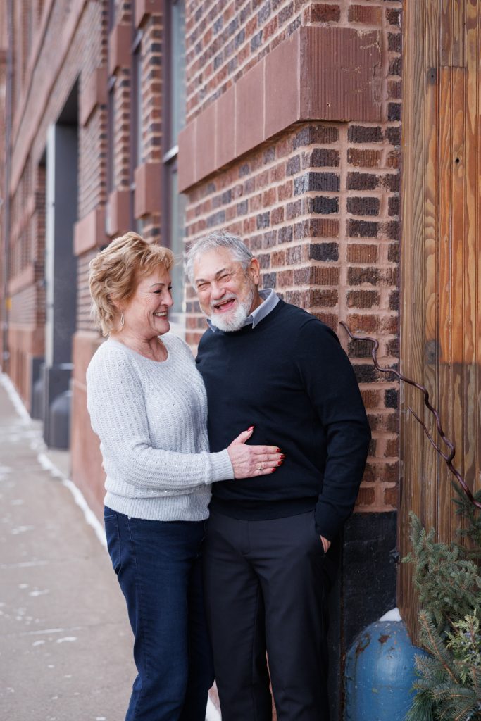 Jane and Bruce smiling together in winter attire, standing against a brick wall in Duluth, Minnesota, capturing a joyful moment from their photography session.