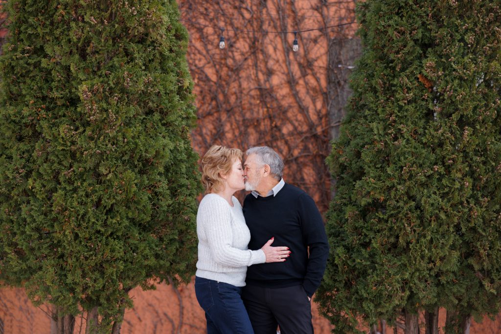 Couple Jane and Bruce sharing a kiss in winter, framed by evergreen trees, in Duluth, Minnesota, showcasing a romantic moment captured by Superior Sunset Studio.