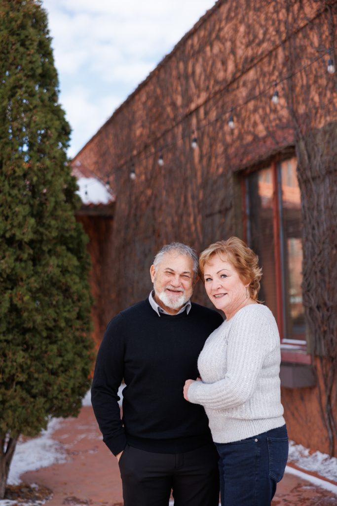 Jane and Bruce smiling together outdoors in Duluth, Minnesota, surrounded by winter scenery and rustic architecture, showcasing a joyful moment captured by Superior Sunset Studio.
