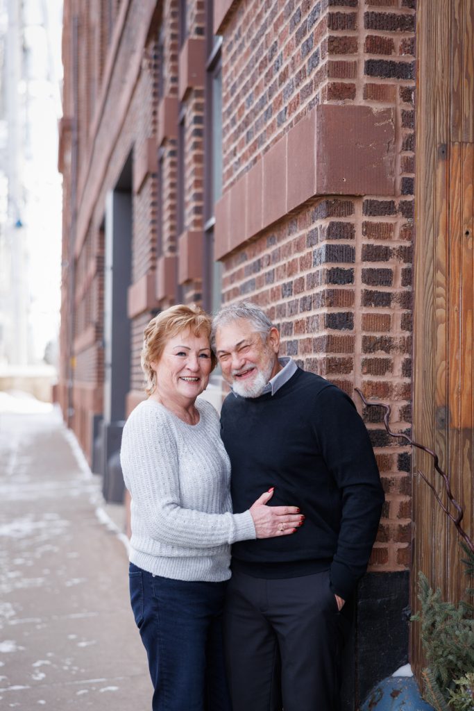 Jane and Bruce smiling together in winter attire, embracing near a brick wall in Duluth, Minnesota, capturing a joyful moment from their photography session.