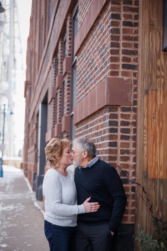 Jane and Bruce sharing a kiss in a winter photography session in Duluth, Minnesota, against a backdrop of brick and wood architecture.