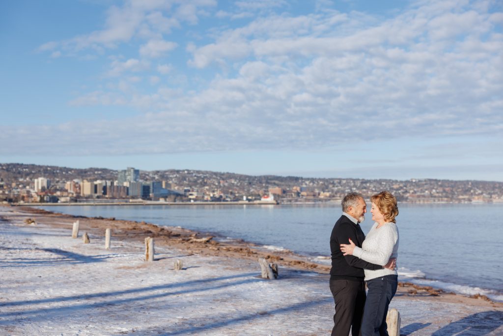 Couple embracing on a snowy Duluth beach, with the city skyline and Lake Superior in the background, capturing a winter portrait session by Superior Sunset Studio.