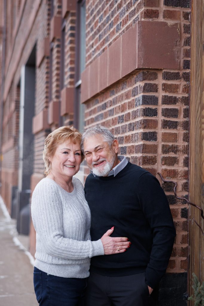 Couple Jane and Bruce smiling together in winter attire against a brick wall in Duluth, Minnesota, celebrating their photography session by Superior Sunset Studio.