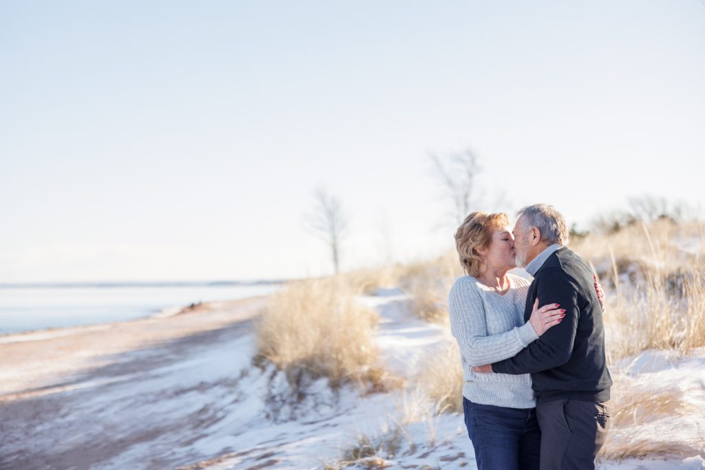 Couple kissing on a winter beach in Duluth, Minnesota, surrounded by snow and tall grass, celebrating love during a photography session by Superior Sunset Studio.