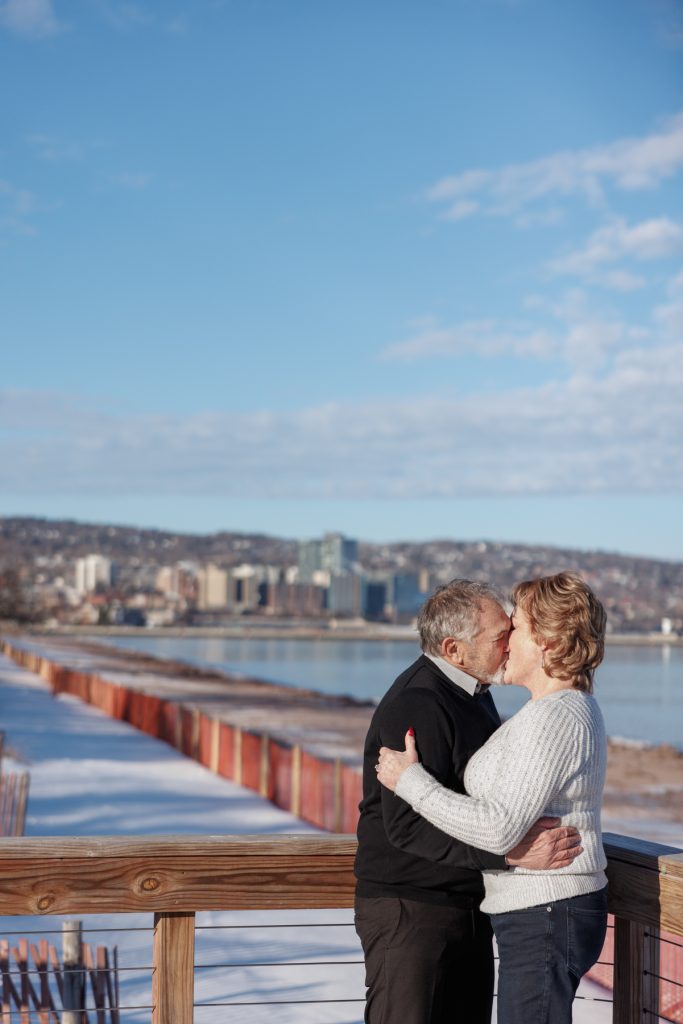 Couple embracing and sharing a kiss on a winter day in Duluth, Minnesota, with a scenic view of the city skyline and frozen lake in the background, showcasing the warmth of their relationship.
