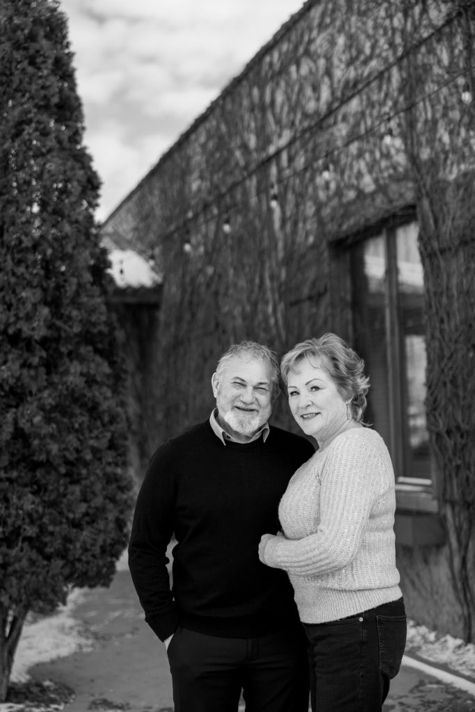 Couple Jane and Bruce smiling together in winter attire, standing in front of a rustic building covered in vines, celebrating their photography session in Duluth, Minnesota.