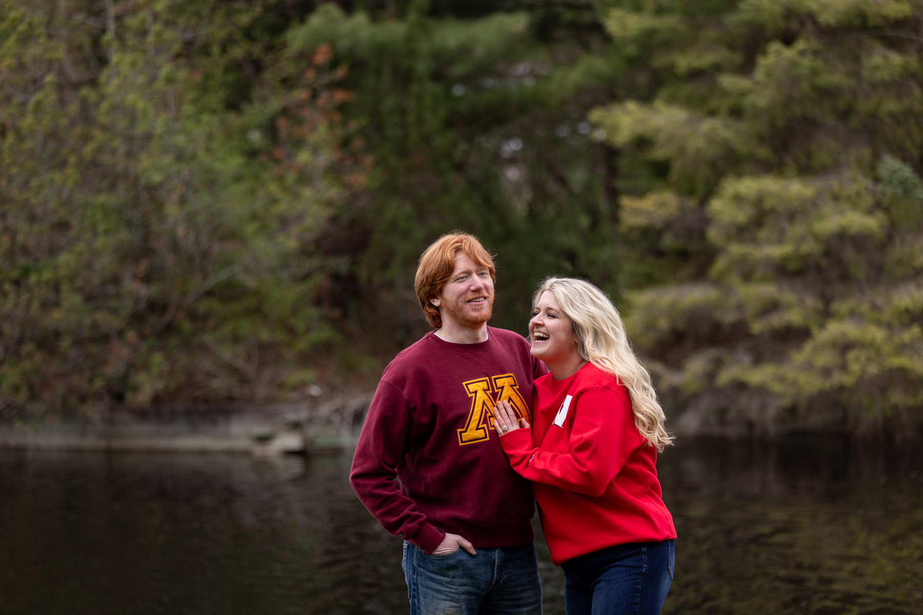 Couple laughing together by a serene lake, featuring a man in a maroon sweatshirt with a university logo and a woman in a red hoodie, capturing a joyful engagement moment in nature.