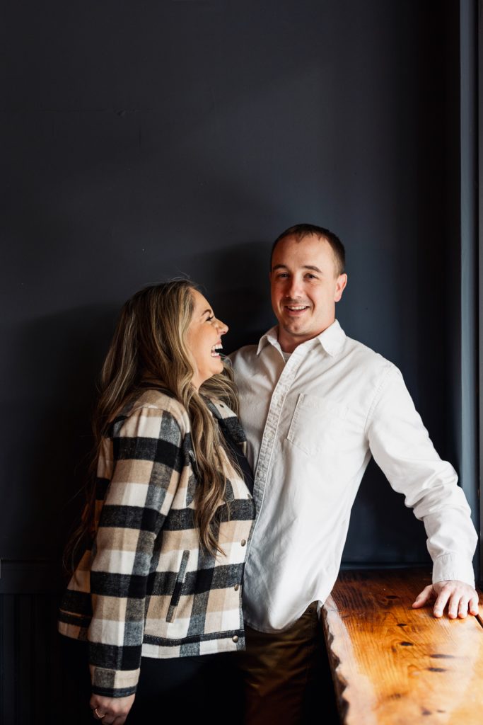 Bianca and Nick smiling together during their engagement session in Hayward, WI, showcasing a cozy indoor setting with a dark background and wooden bar.