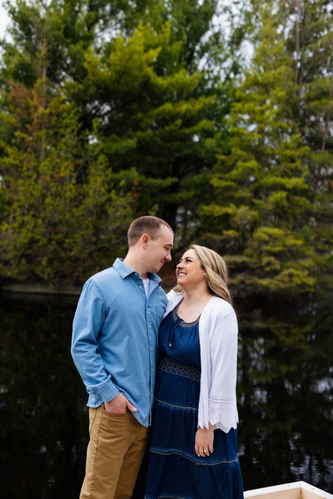 Bianca and Nick smiling at each other during their engagement session in Hayward, WI, surrounded by lush greenery and a tranquil water backdrop.