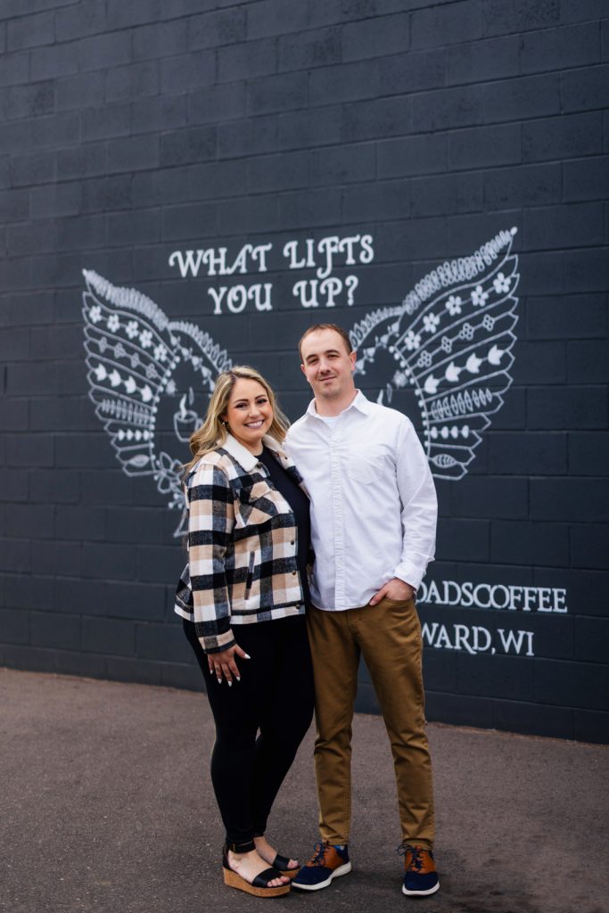 Bianca and Nick posing in front of a mural with angel wings and the text "WHAT LIFTS YOU UP?" during their engagement session in Hayward, WI.