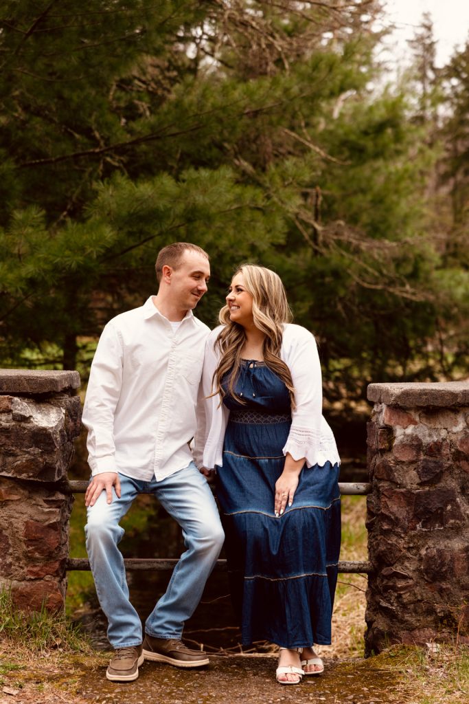 Bianca and Nick sitting on a stone wall surrounded by greenery, smiling at each other during their engagement session in Hayward, WI.