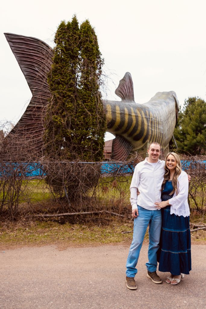 Bianca and Nick posing together in front of a large fish statue in Hayward, WI, showcasing their engagement session captured by Superior Sunset Studio.