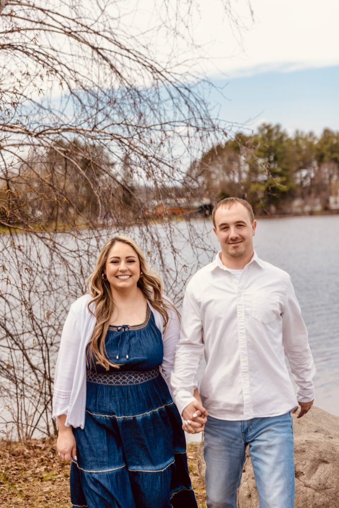 Bianca and Nick holding hands by the water during their engagement session in Hayward, WI, with a scenic backdrop of trees and a lake.