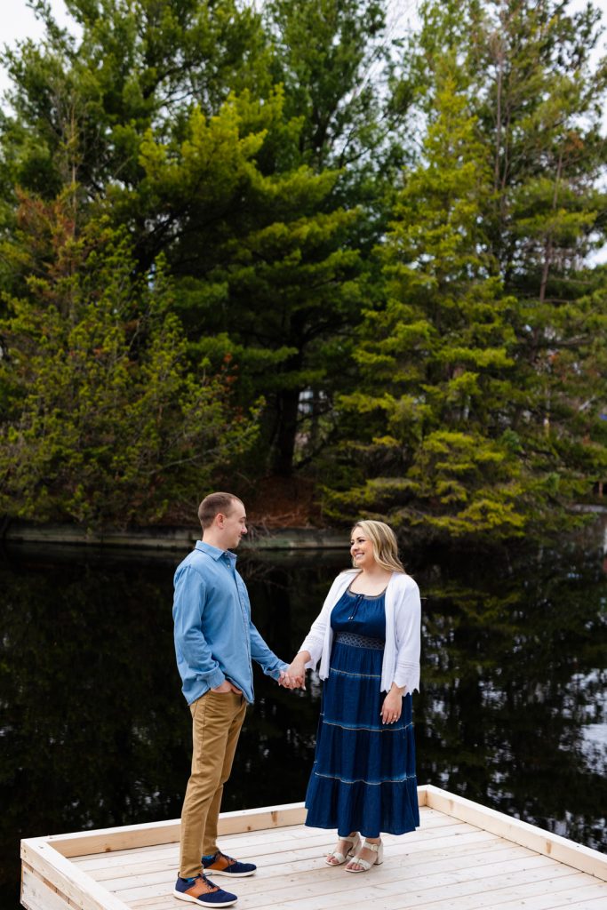 Bianca and Nick holding hands on a wooden platform by a serene lake, surrounded by lush green trees, during their engagement session in Hayward, WI.